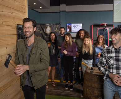 A group of people are gathered in an indoor venue engaged in an axe-throwing activity. One man in the foreground is holding an axe, preparing to throw it at a wooden target on the wall. The group behind him is smiling and appears to be enjoying the event, with some holding drinks. The environment is casual, suggesting a social or recreational setting. Indoor group axe-throwing event with people having fun in a casual venue setting.