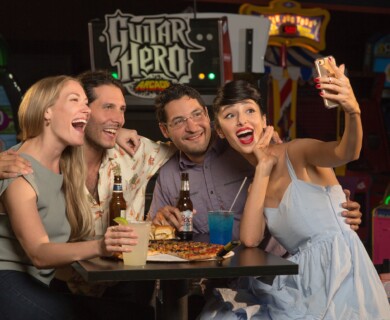 A group of four people is sitting at a table in an arcade, enjoying drinks and food, including a pizza. They are taking a selfie together, appearing happy and engaged in a recreational setting. A "Guitar Hero" arcade game is visible in the background. Group of four taking a selfie at an arcade table with pizza and drinks; Guitar Hero nearby.