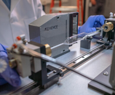 A person wearing blue gloves is working with a precision measuring instrument in a laboratory setting. The equipment, marked with the brand "Keyence," is handling a transparent cylindrical object on a metal rail system. Person with blue gloves using Keyence instrument on transparent object in lab.