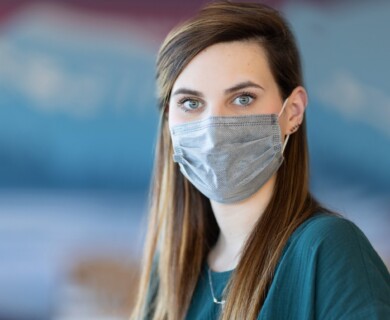 A person with long brown hair is wearing a grey face mask and a green top. The background is blurred with a mix of blue and neutral tones. Person with long brown hair wears a grey mask and green top; blurred blue background.