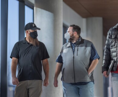 Three masked people walk indoors by windows, engaged in conversation.