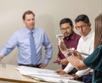 A group of four people engage in a meeting at a table covered with architectural plans. One person holds plastic pipe components, possibly discussing or demonstrating a concept. The setting appears to be professional, likely in an office environment. Meeting with four people discussing architectural plans and pipe components at a table.