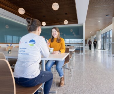Two people converse at a table in a modern cafeteria with blue walls and large windows.