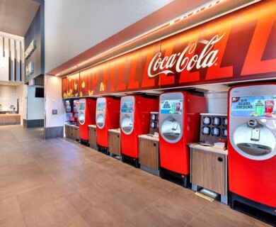 A modern beverage station features several red Coca-Cola Freestyle machines lined up against a wall. The machines allow for various drink customizations, positioned under a large Coca-Cola branded sign. The area is spacious with a counter visible in the background, likely part of a food service area. Modern beverage station with several red Coca-Cola Freestyle machines against a sign.
