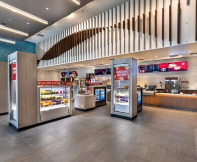 The image shows a modern, well-lit fast-food restaurant interior featuring multiple digital menu boards above the service counter. The space includes various snack display cases and refrigerated sections with beverages. The ceiling has a contemporary design with vertical wooden slats, and the flooring is tiled in a sleek, neutral color. Modern fast-food restaurant interior with digital menu boards, snack displays, and tiled flooring.