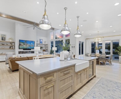 A spacious, modern open-concept living area features a kitchen with a large island, hanging pendant lights, and light wood cabinetry. The adjacent living room includes a brown leather sofa in front of a mounted flat-screen TV and shelves. Large windows provide natural light, and a round dining table is situated near sliding glass doors. The space is decorated in neutral tones with contemporary furnishings and decor. Spacious modern living area with kitchen island, pendant lights, leather sofa, and natural light.