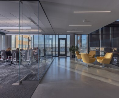 Modern office with glass-walled conference room, people seated, and bright waiting area with chairs.
