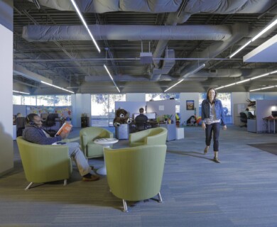 Modern open office with green chairs, workstations, large windows, and exposed ceiling ducts.