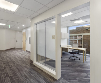 A modern office hallway featuring a glass-walled office space with a desk and chair inside. The hallway and office have ceiling panels and are illuminated by soft lighting. The office is adjacent to a brick exterior wall with large windows. Modern office hallway with glass-walled office, desk, chair, ceiling panels, soft lighting.