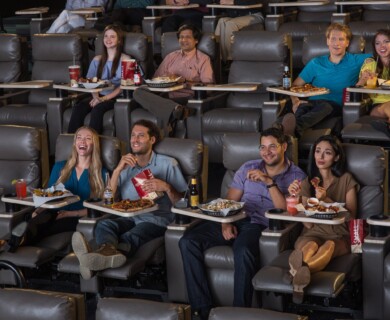 A group of people is seated in a modern movie theater featuring comfortable reclining seats. They are enjoying various snacks and drinks, such as popcorn, pizza, and beverages, while watching a film. The atmosphere appears casual and relaxed. Group in modern theater with reclining seats, enjoying snacks and drinks while watching a film.
