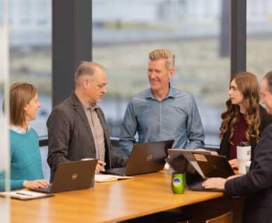 Five people discussing at an office table with laptops and notebooks visible.