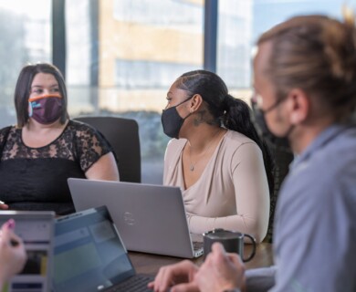 People in face masks sit around a table with laptops in an office meeting.
