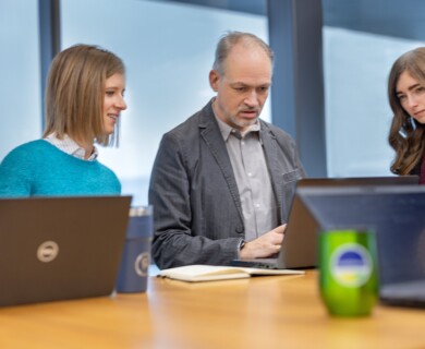 Three individuals are gathered around a table in an office setting, engaged in conversation while using laptops. One person takes notes, and a green mug is visible on the table. Three people discussing around a table with laptops in an office, one taking notes, green mug visible.