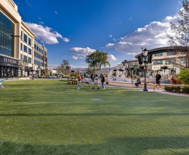 A vibrant outdoor shopping area features a large grassy area with people walking and relaxing. Several stores, including Sephora, are visible along a building to the left, and a fountain area is present in the background under a clear blue sky with scattered clouds. Outdoor shopping area, grassy space, people, Sephora to left, fountain under blue sky with clouds.