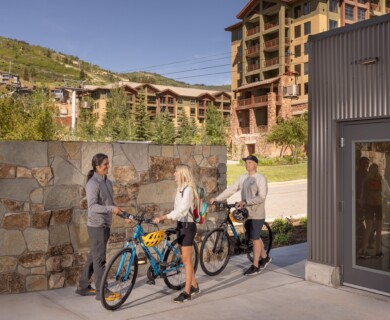 Three people with bicycles near a stone wall and building, with mountains in the background.