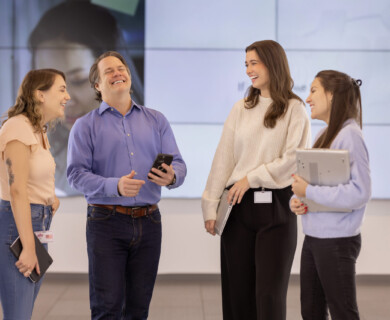 Four people conversing indoors; one holds a smartphone, another a tablet, big screen behind.