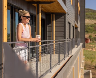 Person in sunglasses on balcony with cup, modern building, greenery, hills, clear sky.