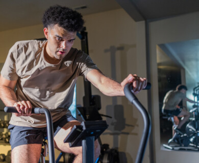 A person is exercising on a stationary bike in a gym setting. The individual is wearing a light-colored T-shirt and dark shorts. There is a large mirror in the background and various gym equipment is visible. Person exercising on a stationary bike in gym, mirror and equipment in background.