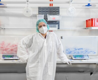 A person in protective clothing, including a lab coat, mask, gloves, and a hairnet, stands in a laboratory or production setting. They are posed in front of workstations with containers filled with colored materials. The environment appears organized, with equipment and storage units visible. Person in protective clothing in lab with containers on workstations.