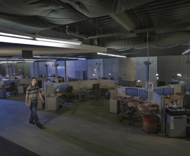 Person walking in a dimly lit office with cubicles, desks, and monitors, under fluorescent lights.