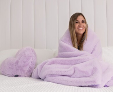 A person wrapped in a soft, lavender-colored blanket sits on a bed, next to a heart-shaped pillow of the same hue. The setting has a light and cozy ambiance with a white, textured headboard and bedding. Person sitting wrapped in a lavender blanket, heart pillow on white bed.