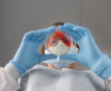 A person wearing protective clothing, a face mask, and blue gloves holds a petri dish with a red substance, examining it closely. Person in protective gear examines a petri dish with red substance using blue gloves.