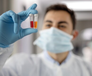 A person wearing a face mask and lab coat holds up two small vials containing red and orange liquids, while wearing blue gloves. Person in lab coat and mask holds vials with red and orange liquids, wearing blue gloves.