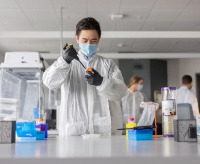A person wearing a mask, gloves, and a protective gown is working with laboratory equipment in a lab setting. The laboratory is equipped with various scientific tools and materials, and other individuals can be seen in the background engaged in similar activities. Person in protective gear using lab equipment, with others in background in a lab setting.