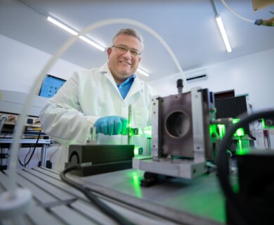 A scientist wearing a lab coat and gloves operates equipment in a laboratory setting, with green light illuminating parts of the machinery. The environment is well-lit and technical, suggesting a research or experimental focus. Scientist in lab coat and gloves operates illuminated equipment in a well-lit laboratory.