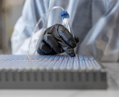A person wearing a black glove is holding a device with a needle-like tip, working on a surface covered with multiple small wells. The person appears to be in a laboratory setting, wearing a protective gown. Person in lab working with needle-tipped device on multi-well surface, wearing black glove.