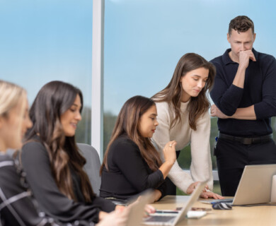 A group of five people gathered around a table with laptops in a meeting room. Some are seated while others stand, focusing on a laptop screen, appearing engaged in a discussion. Large windows in the background show a clear sky. Five people in a meeting room, focused on laptops, discuss around a table with large windows.