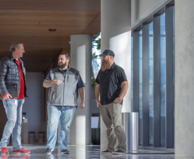 Three men walking in a modern corridor with windows and concrete pillars, engaged in conversation.