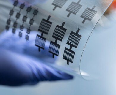 A flexible transparent sheet is displayed, featuring a pattern of printed electronic circuits. A gloved hand is holding the sheet, highlighting its bendability. Flexible transparent sheet with printed electronic circuits held by a gloved hand.