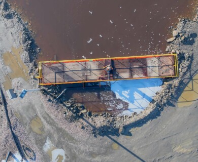 An aerial view of a construction site featuring a narrow, elevated platform or bridge with safety railings. The platform extends over a muddy, brown waterway, surrounded by rocky and uneven terrain. A worker in protective gear is positioned on the bridge, engaged in maintenance or construction tasks. The area shows signs of industrial activity with equipment visible. Aerial view of a construction site with a worker on a platform over muddy water.