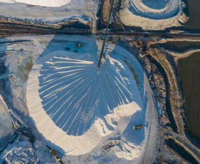 An aerial view of an industrial site featuring a large circular mound of material with machinery operating nearby. The area is bordered by water bodies and intersected by rail tracks, displaying a network of pathways and surrounding terrain. Aerial view of industrial site with circular mound, machinery, rail tracks, water bodies, and paths.