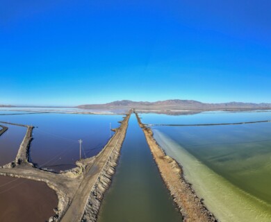 Aerial view of expansive salt flats featuring sections of colorful water in hues of brown, blue, and green, intersected by narrow dirt roads. The landscape is bordered by distant mountain ranges under a clear blue sky. Aerial view of vast salt flats with colorful water sections and dirt roads, distant mountains visible.