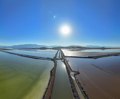 A symmetrical aerial view of expansive salt flats or evaporation ponds, bordered by narrow roads and channels. The landscape is reflected in the water, and distant mountains under a clear blue sky are visible in the background, with the sun positioned low on the horizon. Aerial view of symmetrical salt flats with roads, reflecting mountains and sun on the horizon.