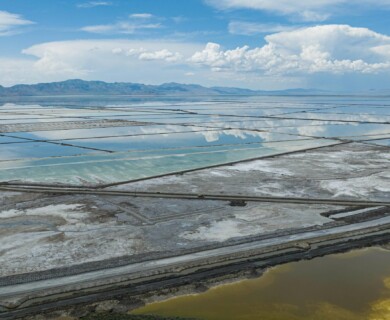 Aerial view of expansive salt flats with reflective water surfaces, bordered by geometric patterns and contrasting colors. Mountains and a cloudy sky are visible in the background. Aerial view of salt flats with reflective water, geometric patterns, mountains, and cloudy sky.