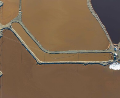 An aerial view of salt evaporation ponds shows contrasting sections of water in shades of brown and green, separated by narrow, linear barriers. The ponds are connected by a system of roads or pathways, with some small structures or equipment visible near the junctions. Aerial view of salt ponds with brown and green water, separated by barriers, roads, and structures.