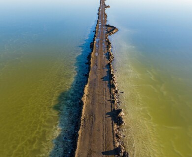 A narrow dirt road extends through a body of water, dividing it into two sections. The water appears varied in hues, with greenish and blue tones visible on either side of the road. Rocks and slight puddles are present along the road’s surface. Narrow dirt road divides greenish-blue water with varied hues and visible rocks, puddles.