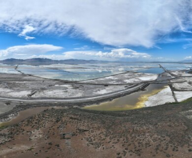 A panoramic view of a landscape featuring a large expanse of salt flats near a body of water, with visible industrial structures in the distance. The foreground includes barren terrain with sparse vegetation, while the background displays mountains under a partly cloudy sky. Panoramic landscape of salt flats with industrial structures, barren terrain, and distant mountains.